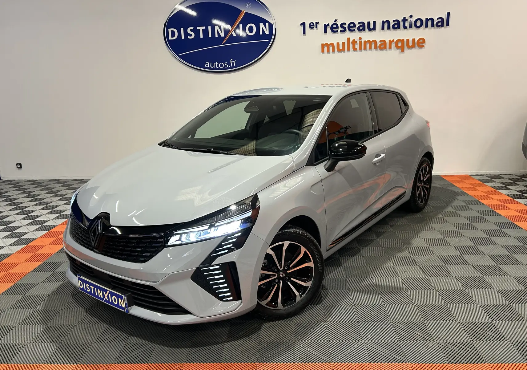 Renault Clio V E-Tech gris Rafale métal en 3/4 avant droit, phares LED allumés et jantes noires bicolores.