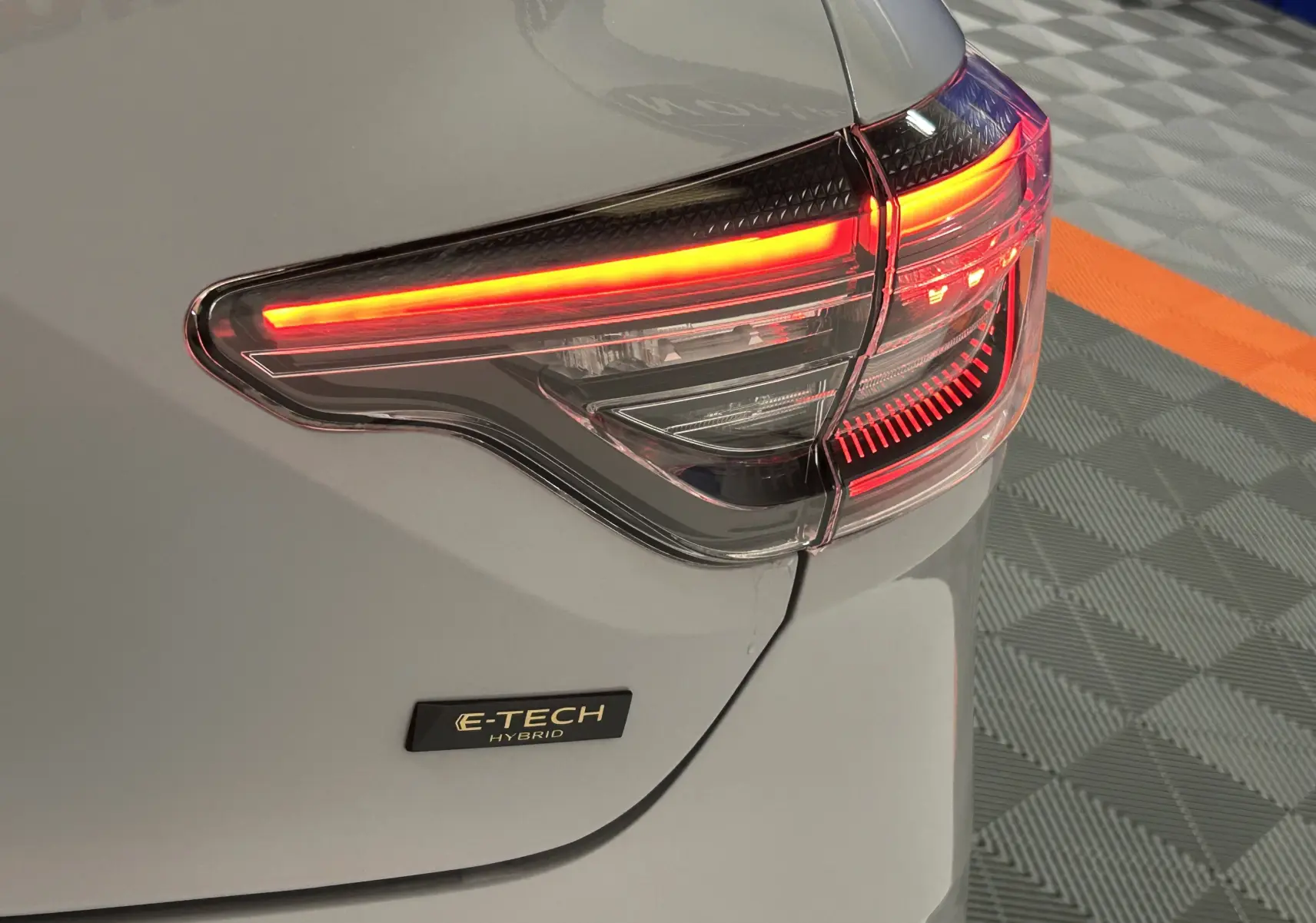 Gros plan sur le feu arrière droit allumé et le badge E-TECH sur la carrosserie gris rafale métal d'une Renault Clio V hybride.