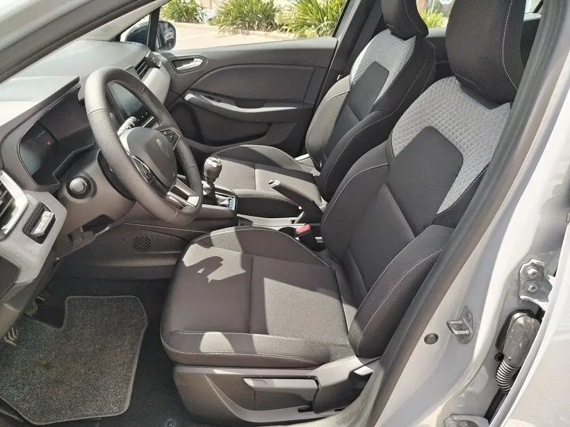 Vue intérieure côté conducteur de la Renault Clio V TCE 90 Evolution 2026, sièges tissu noir et gris, volant multifonction et levier de vitesse manuel.