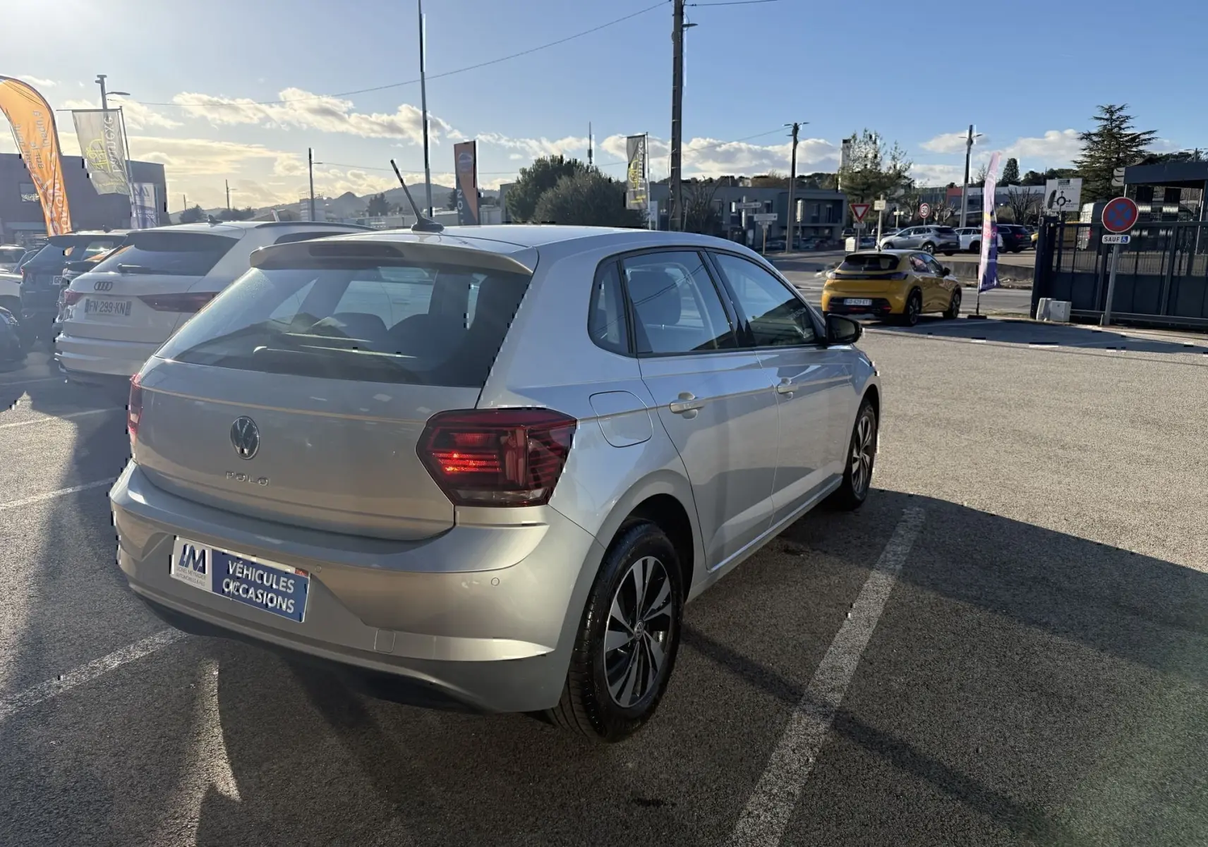 Volkswagen Polo gris métal vue 3/4 arrière droit, avec feux arrière allumés, stationnée en extérieur au soleil.