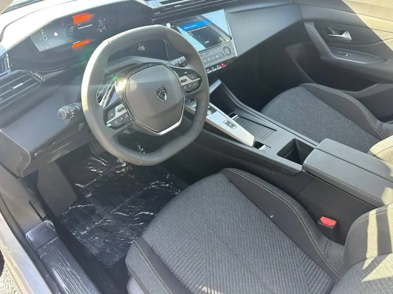 Vue intérieure côté conducteur de la Peugeot 308 2025, volant multifonction noir et sièges tissu gris foncé.