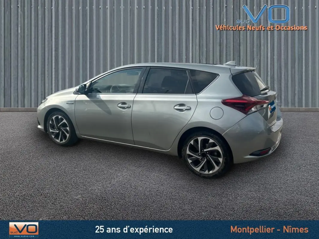 Profil côté gauche d'une Toyota Auris Business Hybride gris clair de 2016 avec jantes alliage 17 pouces sur fond urbain.