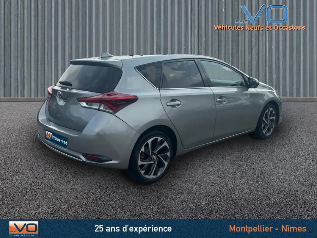 Toyota Auris Business Hybride gris clair vue 3/4 arrière droit avec jantes alliage 17 pouces sur parking.