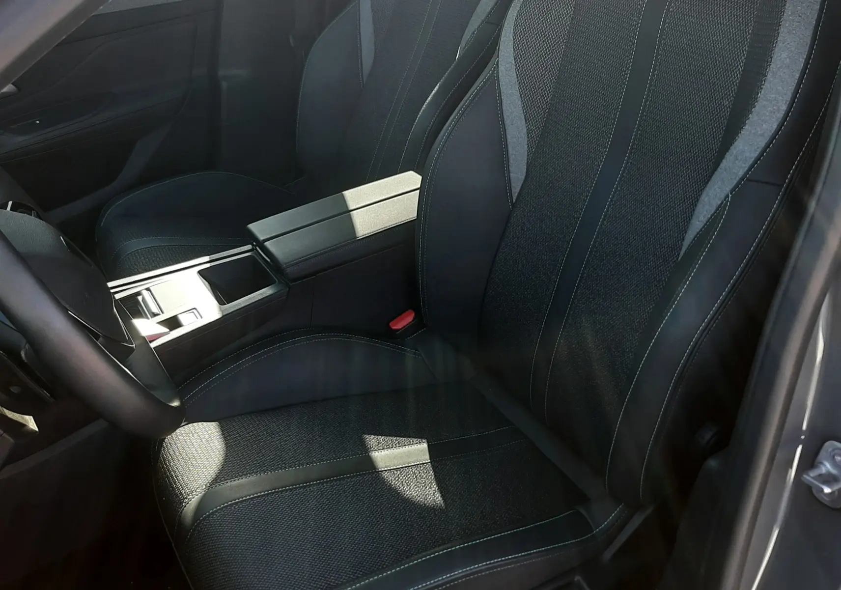 Vue intérieure côté conducteur de la Peugeot 308 SW Hybrid 2025, sièges tissu noir avec surpiqûres claires et console centrale.