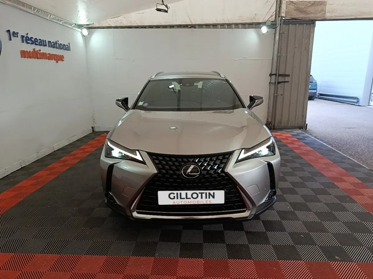 Vue avant d'un Lexus UX 250H gris clair avec calandre noire en losange et phares LED allumés dans un showroom.