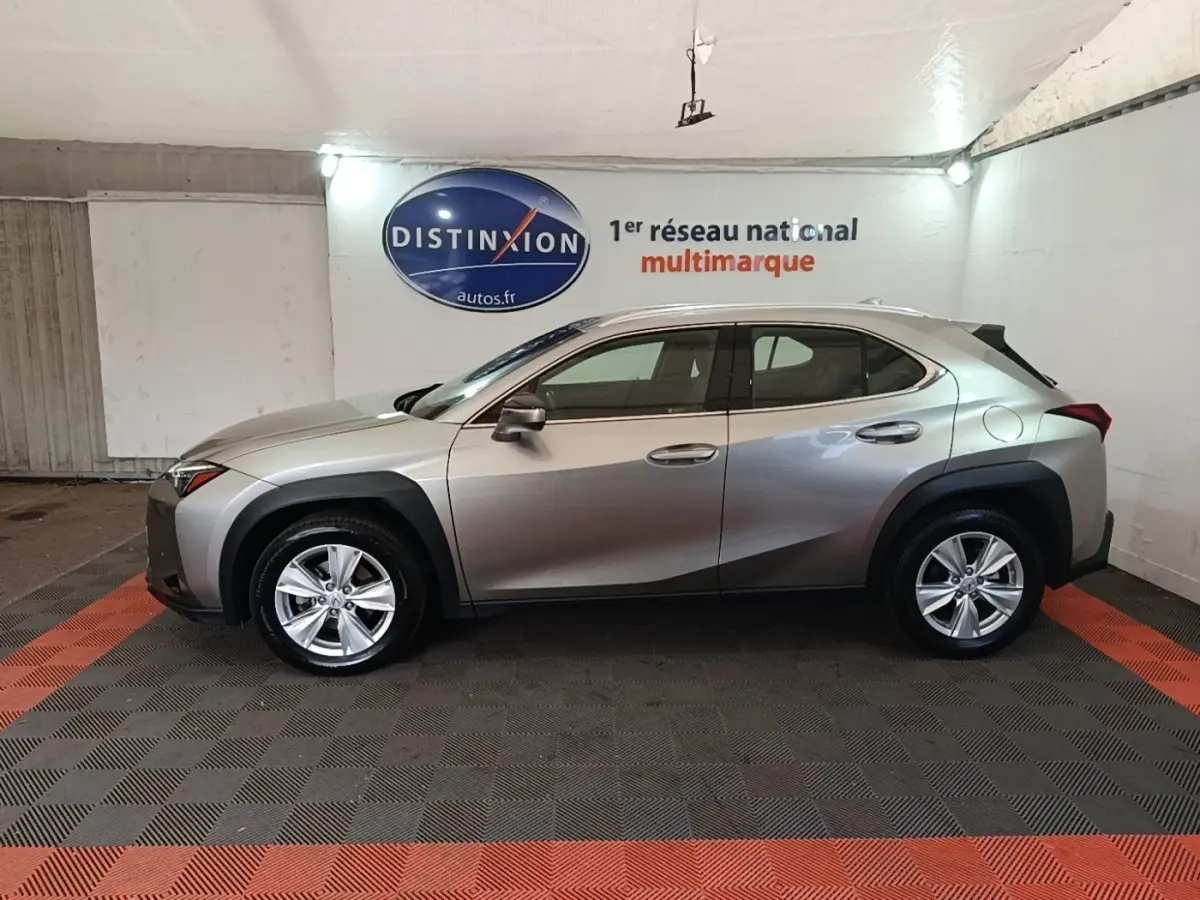 Profil droit d'un Lexus UX 250H gris clair avec protections noires autour des passages de roues, en intérieur showroom.