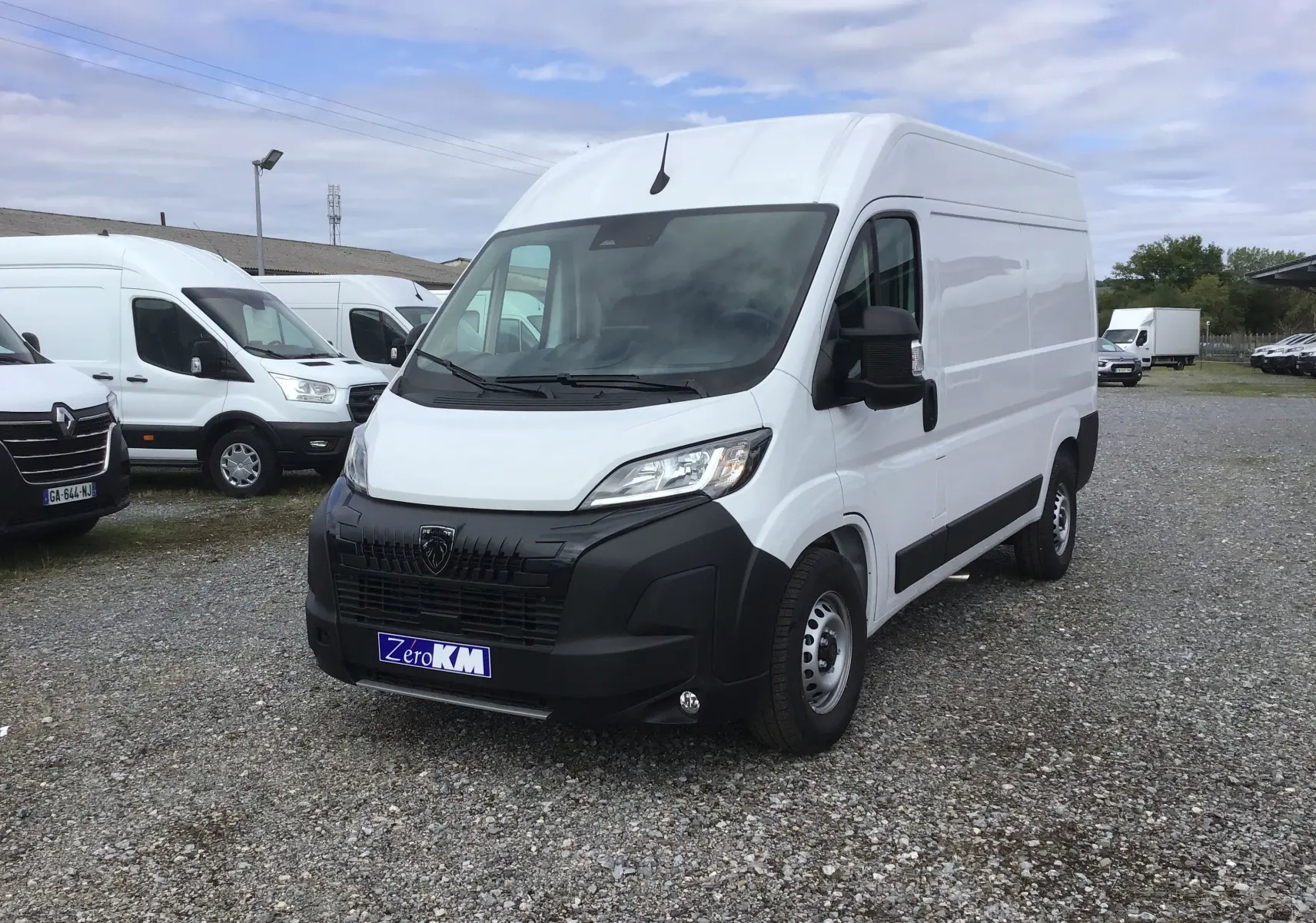 Fourgon Peugeot Boxer blanc en 3/4 avant droit sur parking gravillonné avec plusieurs véhicules utilitaires en arrière-plan.