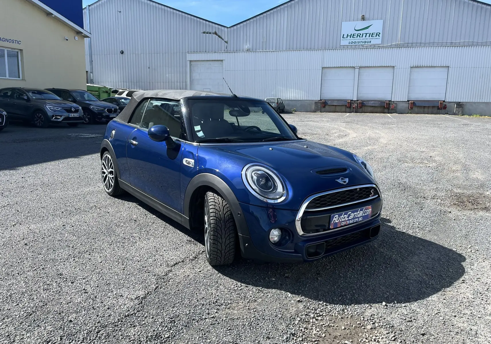Vue 3/4 avant droite d'une MINI Cabriolet bleu avec capote beige et jantes bi-ton sur un parking industriel.