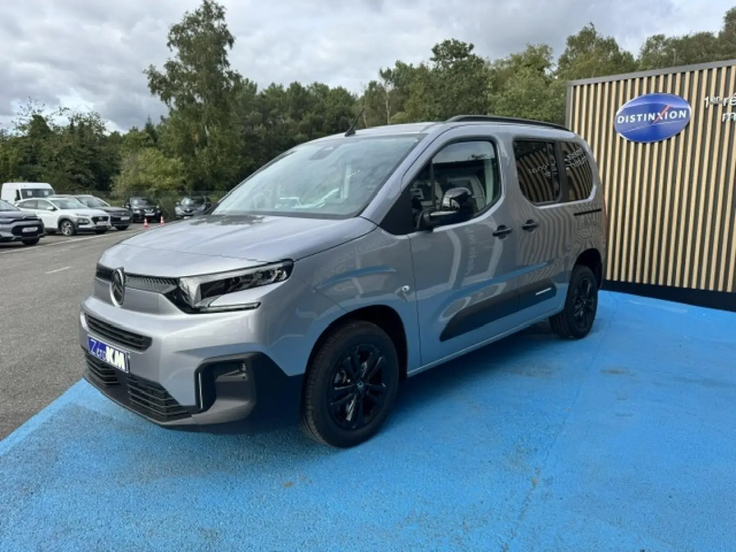 Citroën Berlingo gris acier en 3/4 avant droit, avec jantes noires et barres de toit visibles.