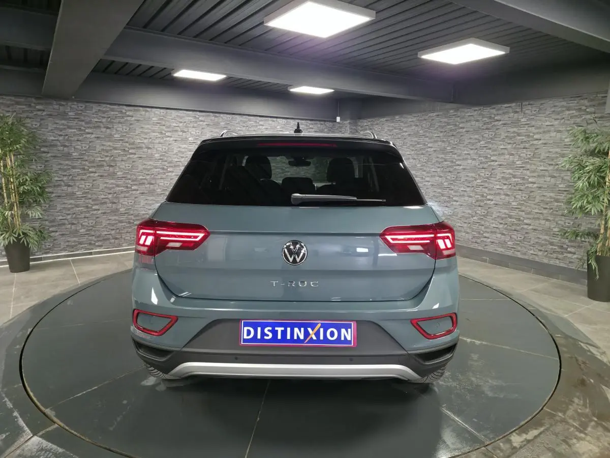 Vue arrière d’un Volkswagen T-Roc bleu métallisé 2025 avec toit noir et feux arrière LED allumés dans un showroom.