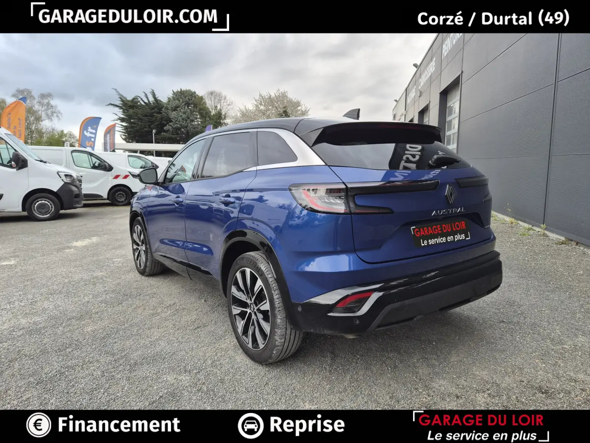 Vue 3/4 arrière droite d'un Renault Austral bleu avec toit noir et jantes alliage, stationné devant un bâtiment gris.