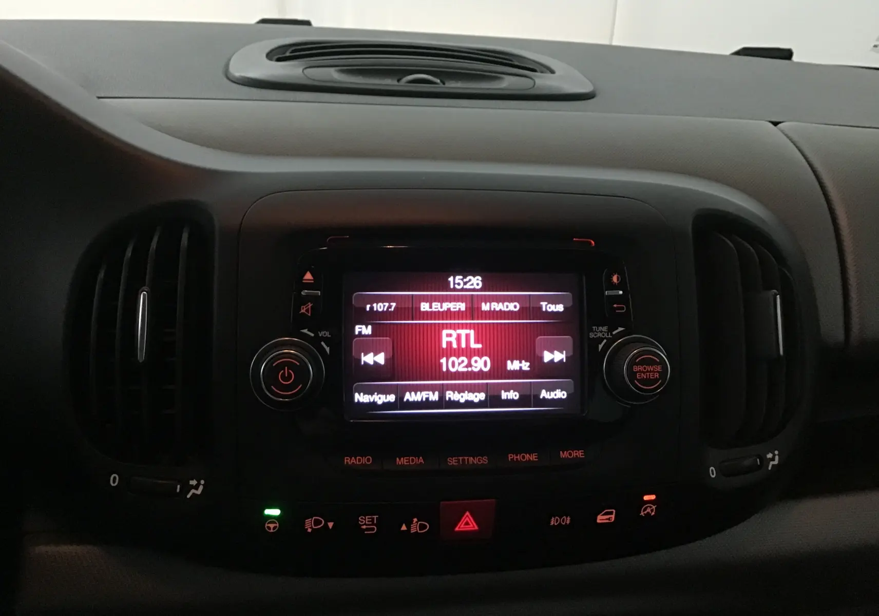 Tableau de bord de Fiat 500L blanc 2015 avec écran central allumé sur radio RTL 102.9 MHz, ambiance intérieure sombre.