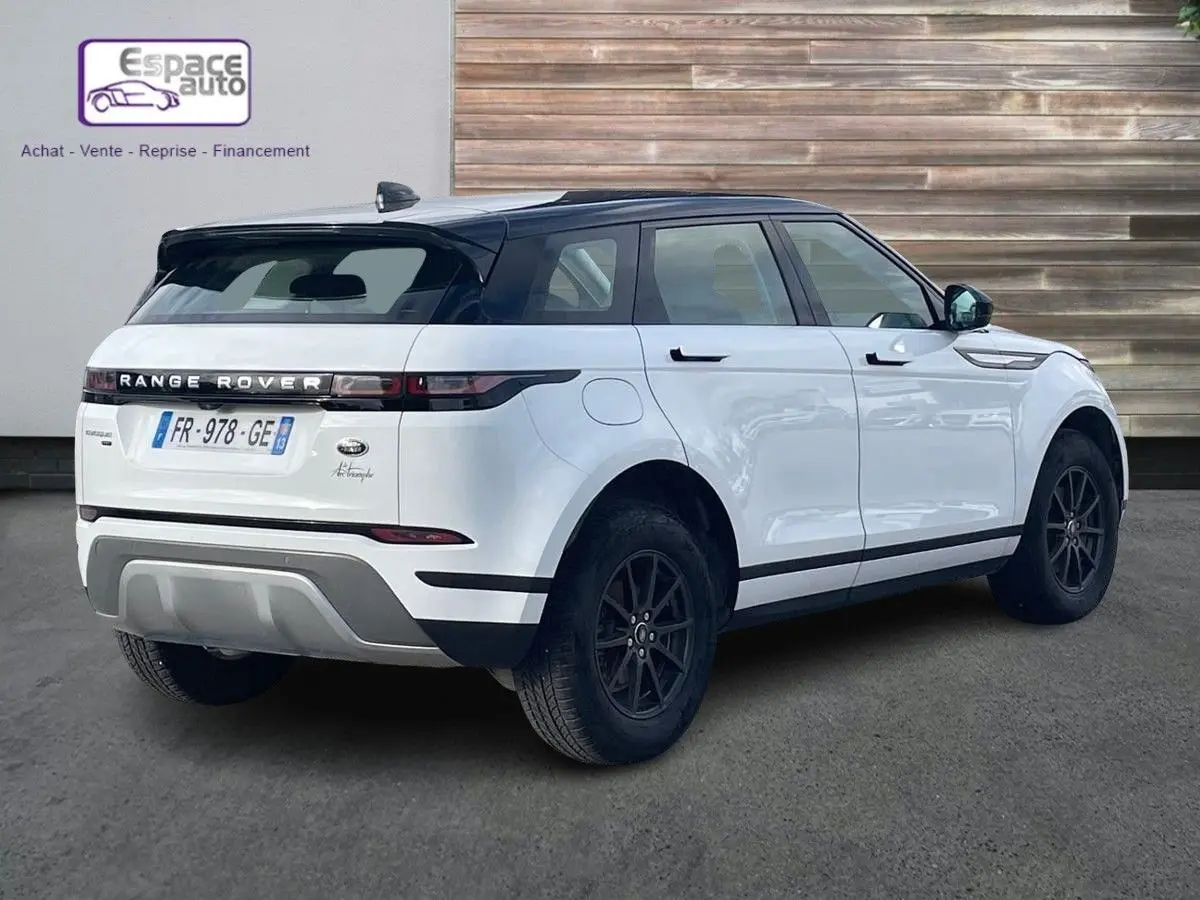Vue 3/4 arrière droite d'un Range Rover Evoque blanc 2020 avec toit noir et jantes foncées, stationné devant un mur en bois.