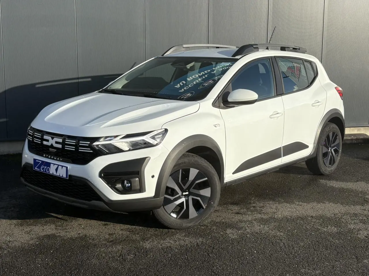 Vue 3/4 avant droite d'une Dacia Sandero Stepway 2024 blanc glacier avec barres de toit et jantes noires.