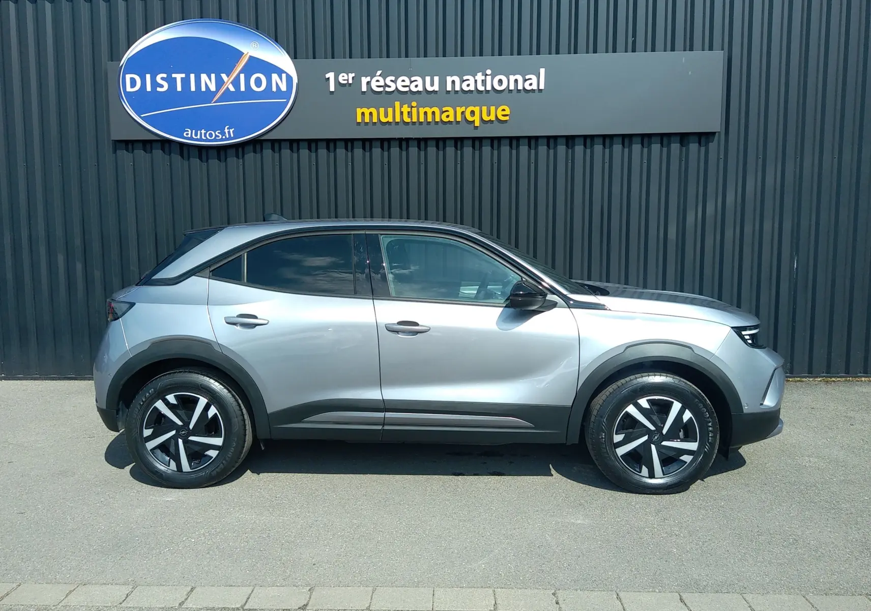 Vue de profil côté gauche d’un Opel Mokka Electric gris 2025, SUV compact avec jantes noires et grises, garé devant un mur sombre.