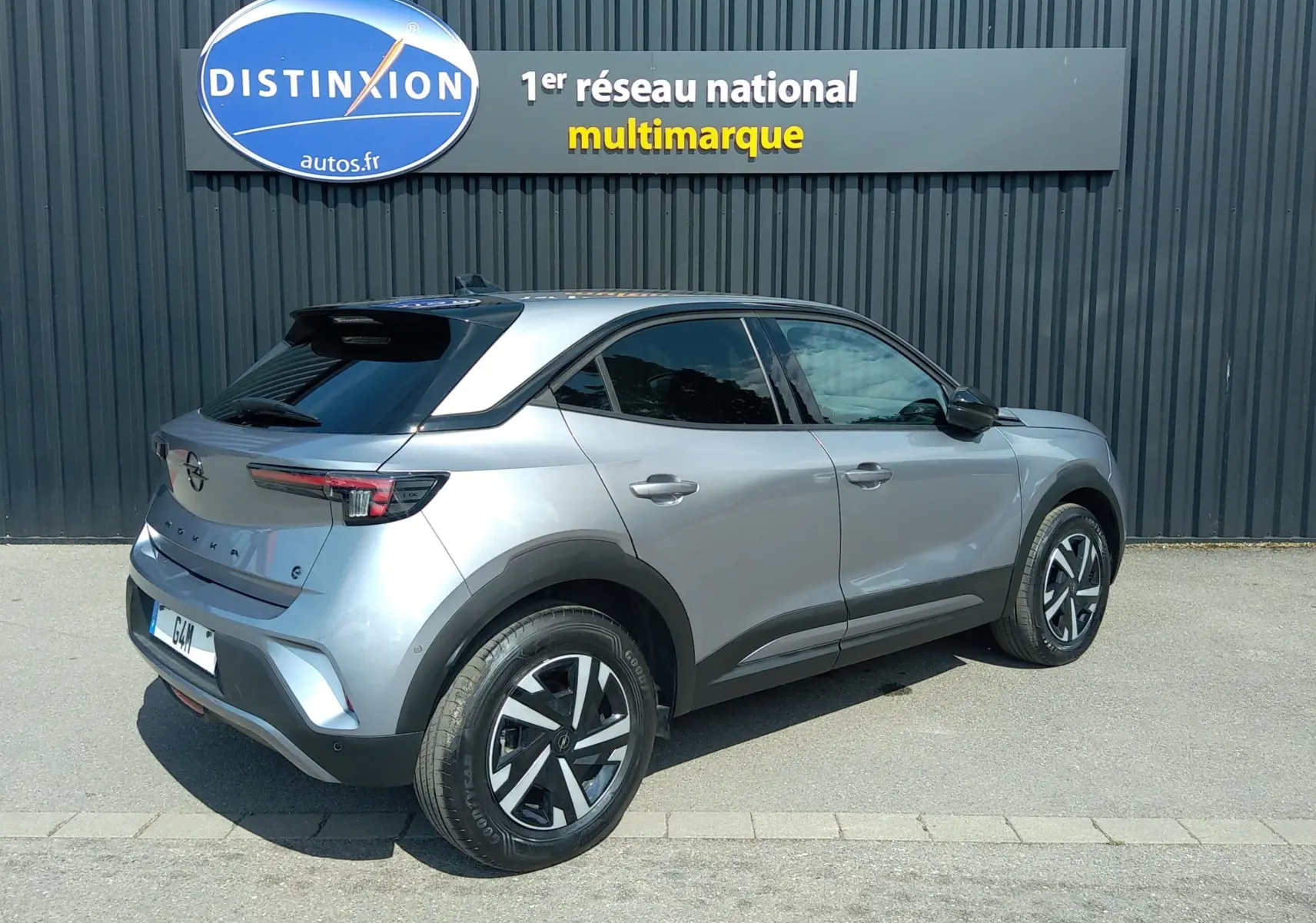 Opel Mokka Electric gris en 3/4 arrière droit, avec toit noir et jantes distinctives, stationné devant un mur gris.
