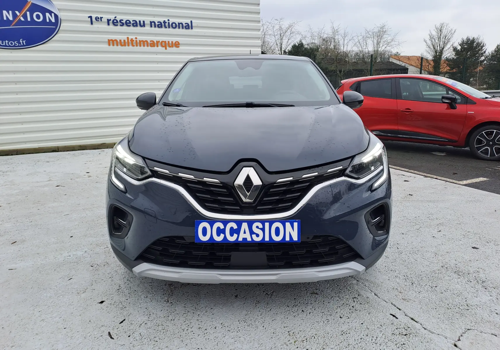 Vue avant d'un Renault Captur bleu marine 2022 avec calandre chromée et phares LED allumés sur parking.