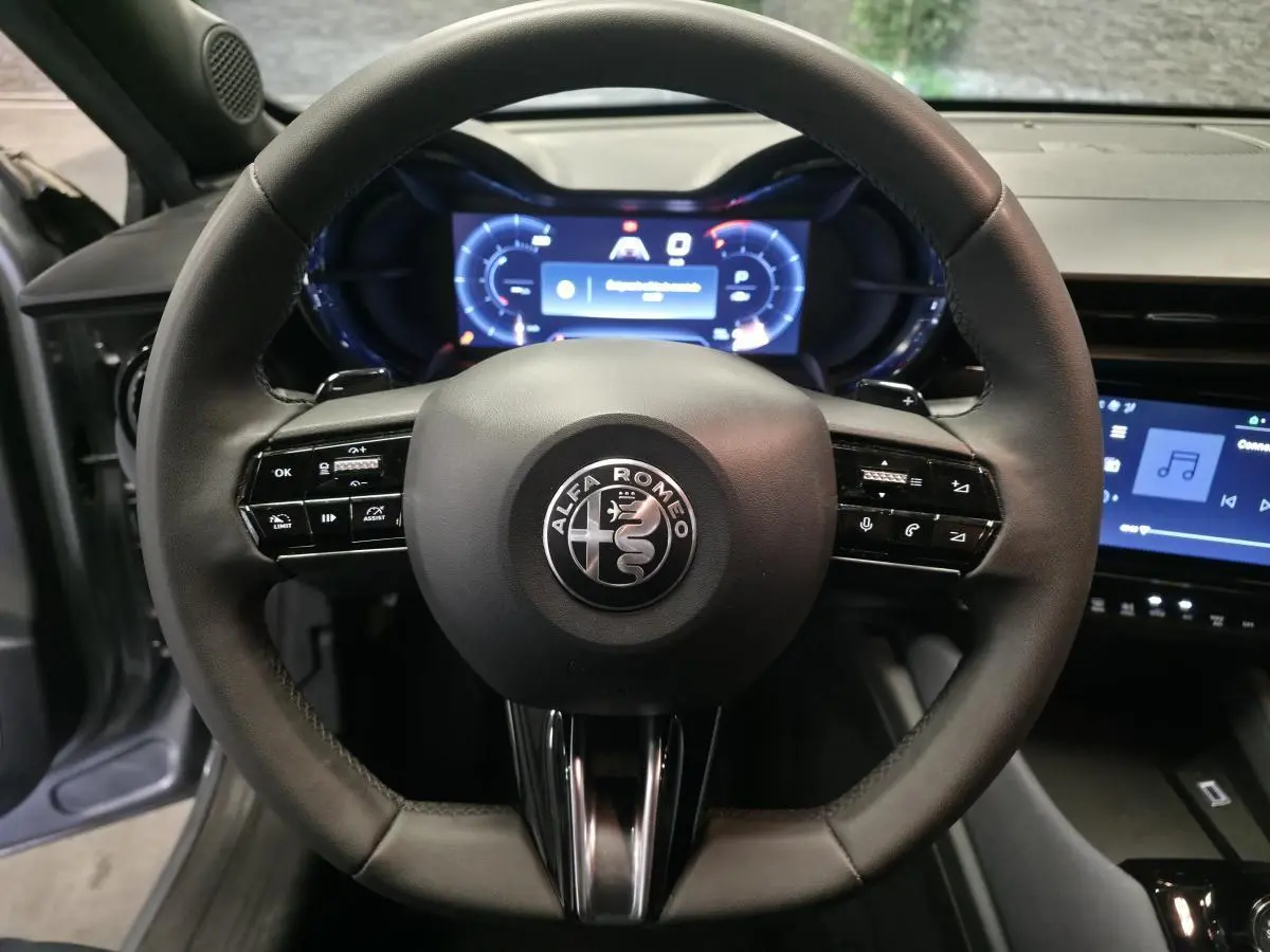 Vue rapprochée du volant noir multifonctions d'une Alfa Romeo Junior 1.2 Ibrida avec tableau de bord digital bleu.