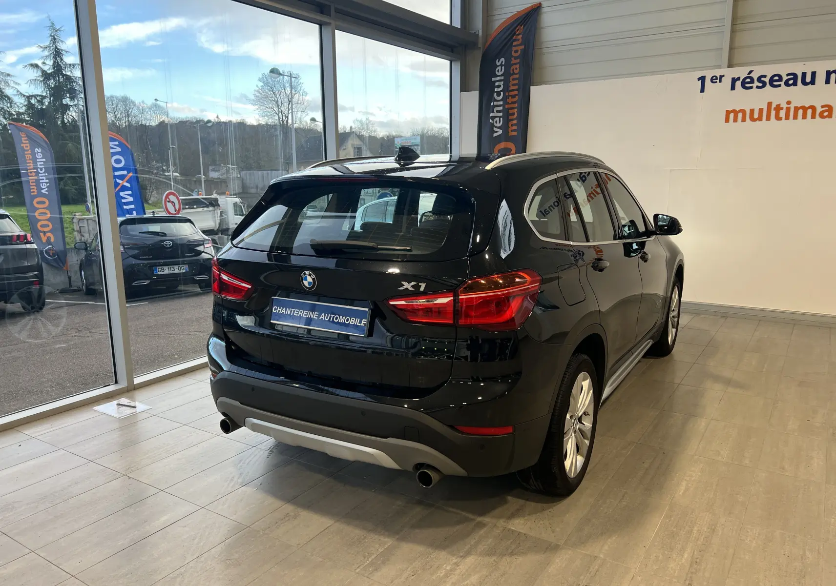 BMW X1 F48 noir saphir, vue 3/4 arrière droit en showroom avec logo X1 et double sortie d’échappement visible.