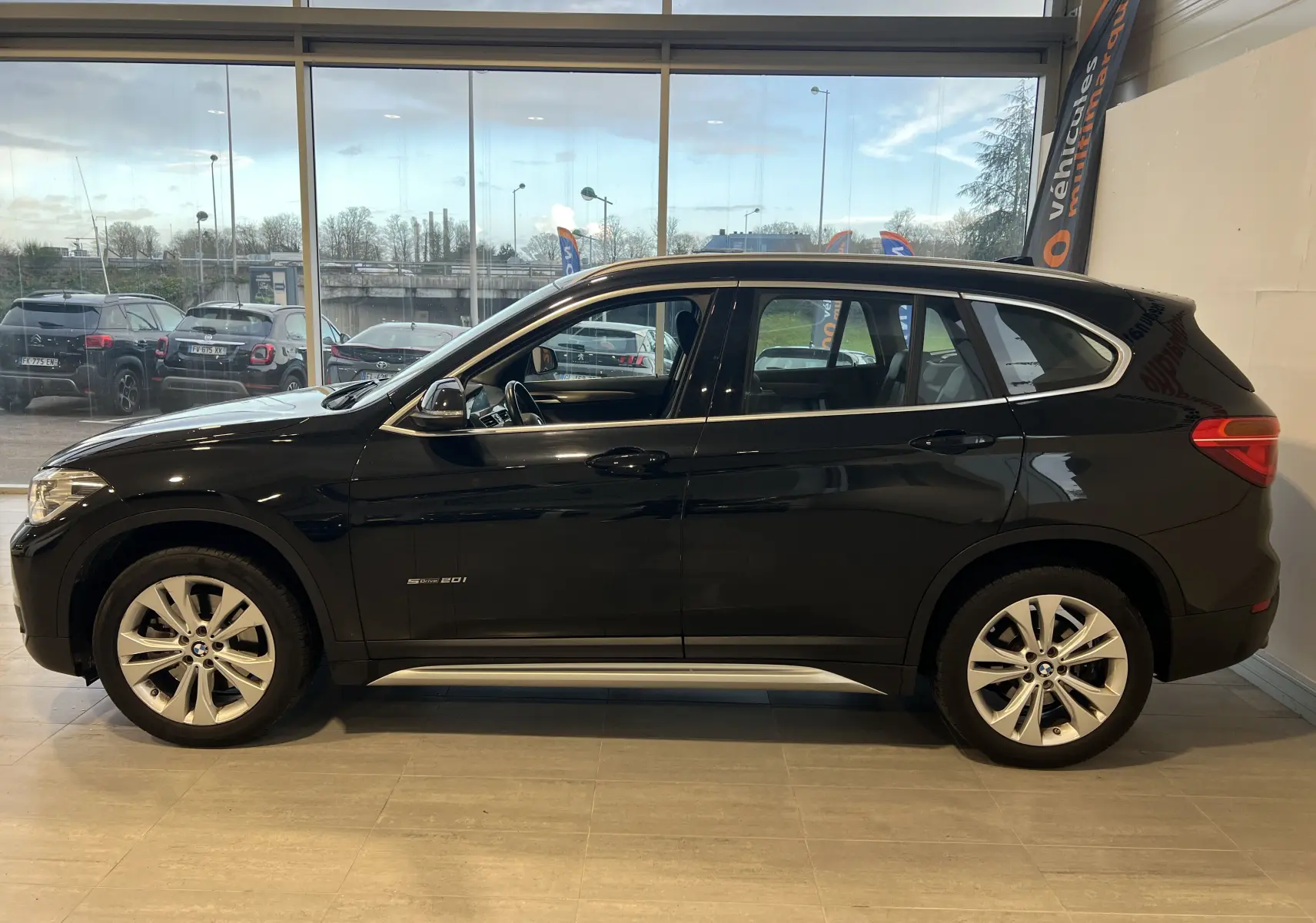 Profil côté gauche d'un BMW X1 F48 noir Saphirschwarz de 2017, finition sDrive16d Lounge, en intérieur showroom.