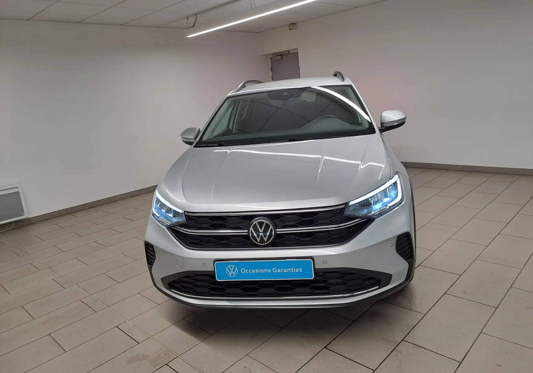 Vue avant d'un Volkswagen Taigo 2025 en peinture métallisée Reflet d'argent avec phares allumés dans un showroom.
