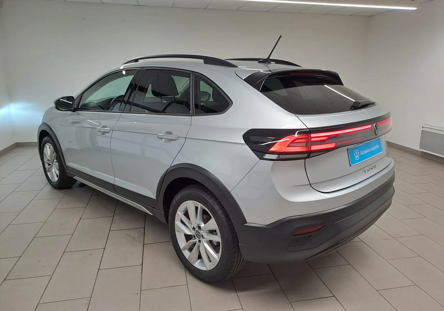 Vue 3/4 arrière droite du Volkswagen Taigo 2025 en peinture métallisée Reflet d'argent avec feux arrière allumés.