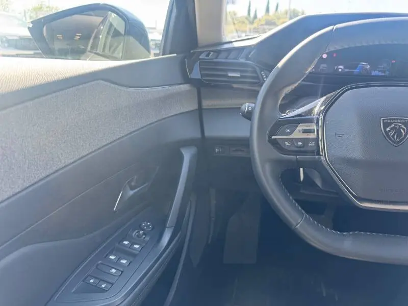 Vue intérieure côté conducteur de la Peugeot 308 Hybrid 145 e-DCS6 Allure 2025, volant multifonctions et commandes vitres électriques.