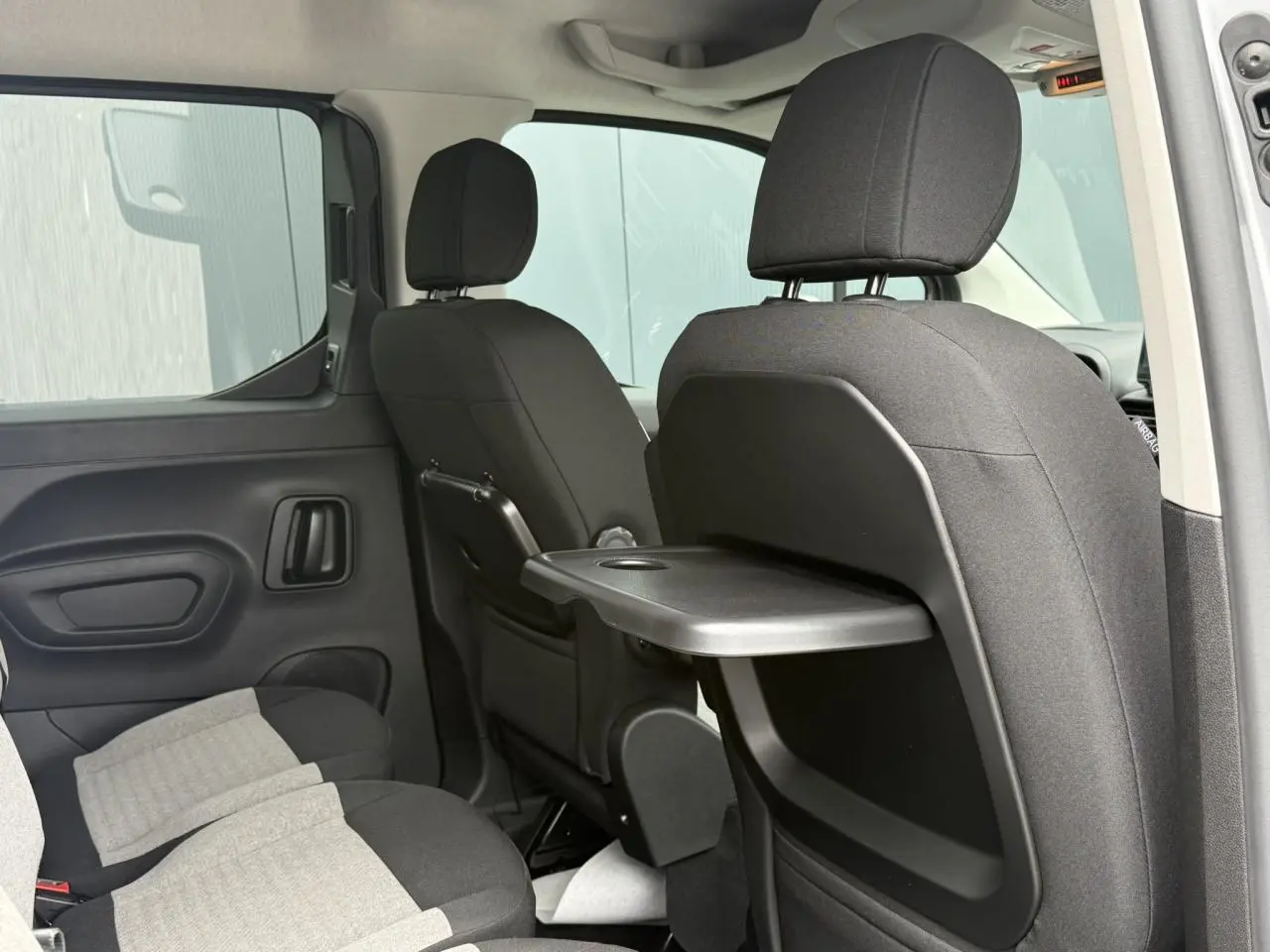 Vue intérieure arrière droite du Citroën Berlingo gris acier montrant les sièges avec tablette rabattable et lunette arrière ouvrante.
