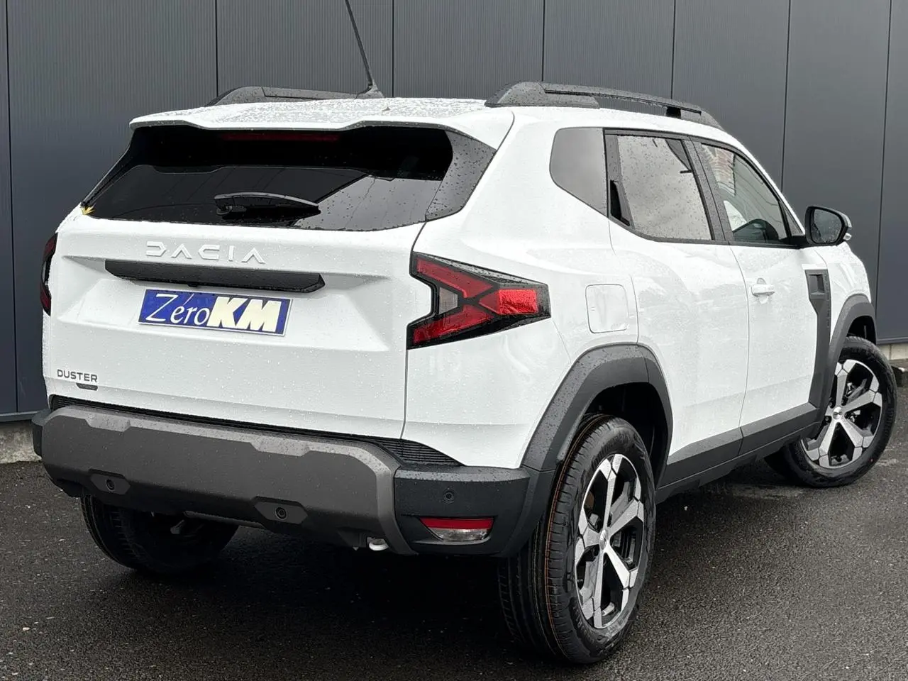 Vue 3/4 arrière droit du Dacia Duster blanc Glacier 2025 avec jantes alliage et barres de toit noires.