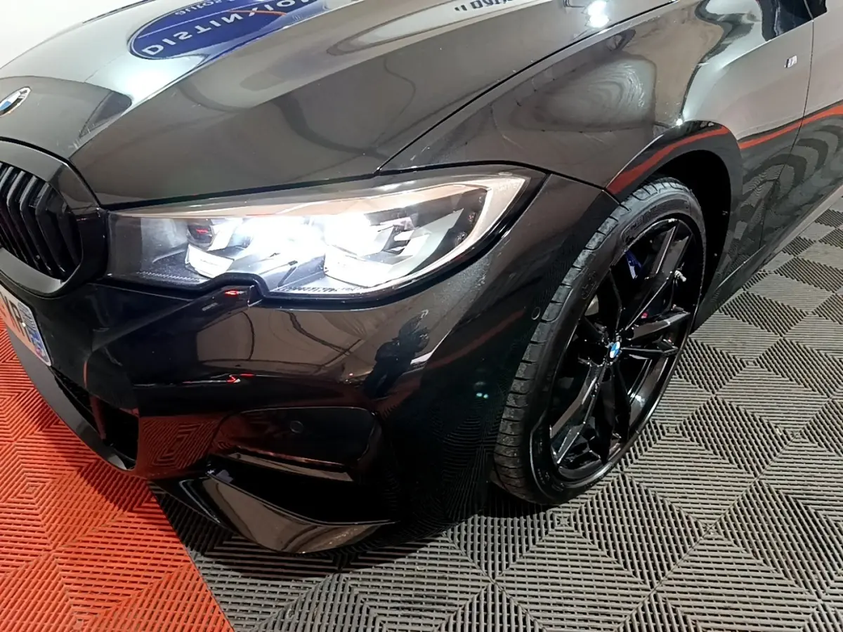 Vue 3/4 avant droit d'une BMW Série 3 318i noire avec jantes noires brillantes et phares LED allumés.