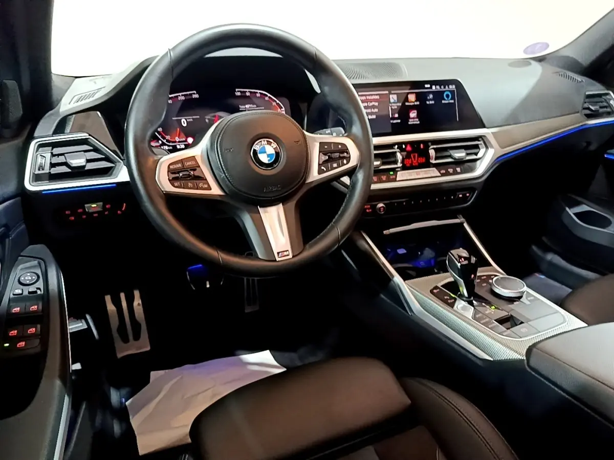 Vue intérieure avant de la BMW Série 3 318i M Sport 2022, volant cuir M, tableau de bord digital et éclairage d'ambiance bleu.
