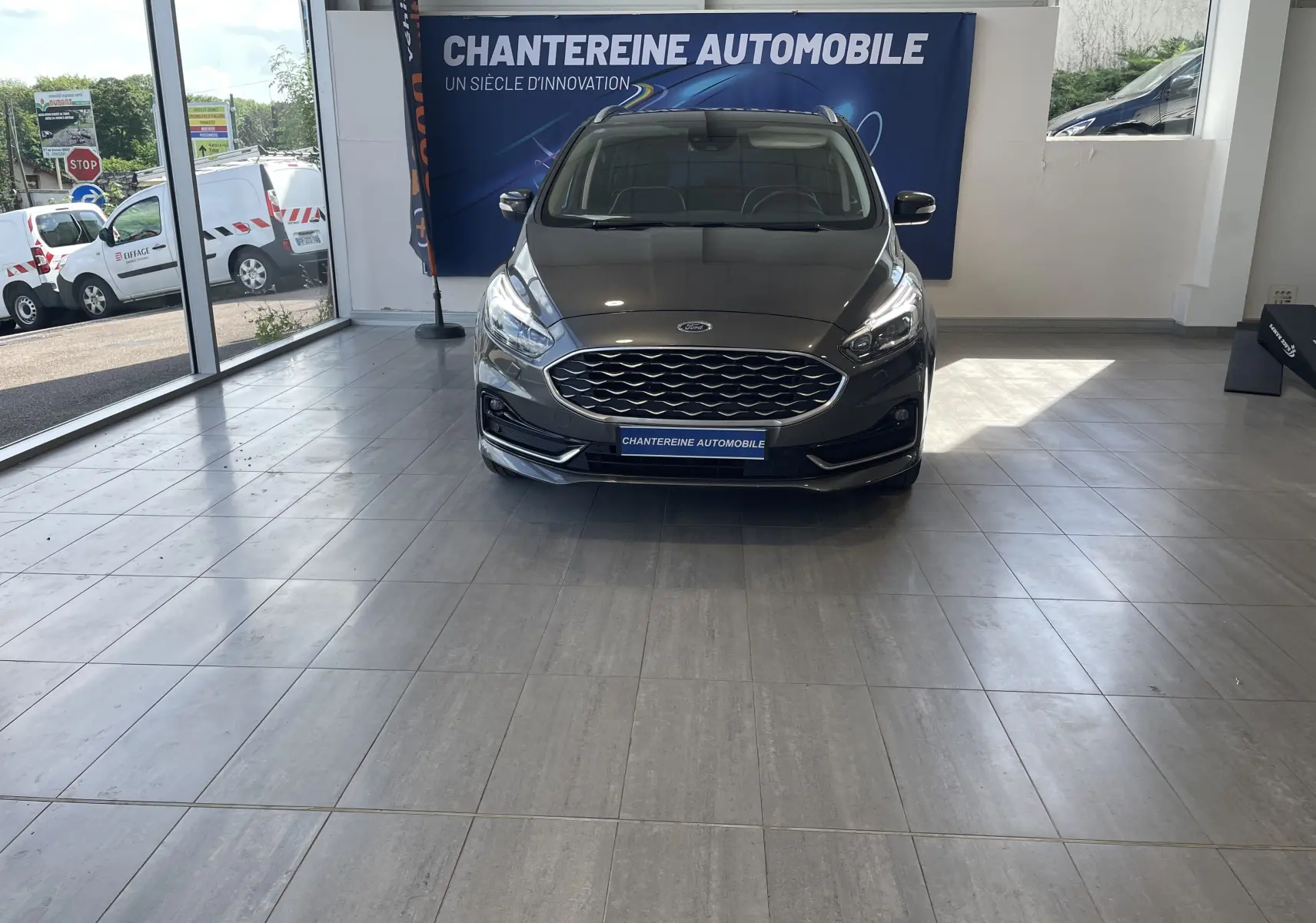 Vue frontale d'un Ford S-MAX Vignale gris magnetic 2022, avec calandre hexagonale et phares LED allumés en showroom.