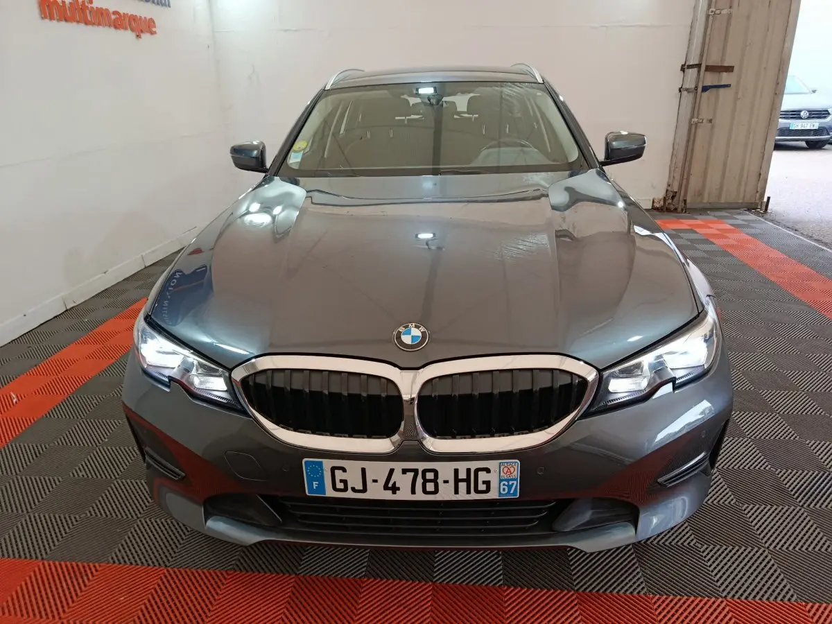 BMW Série 3 316d gris clair vue de face en intérieur, avec calandre double haricot et phares LED allumés.