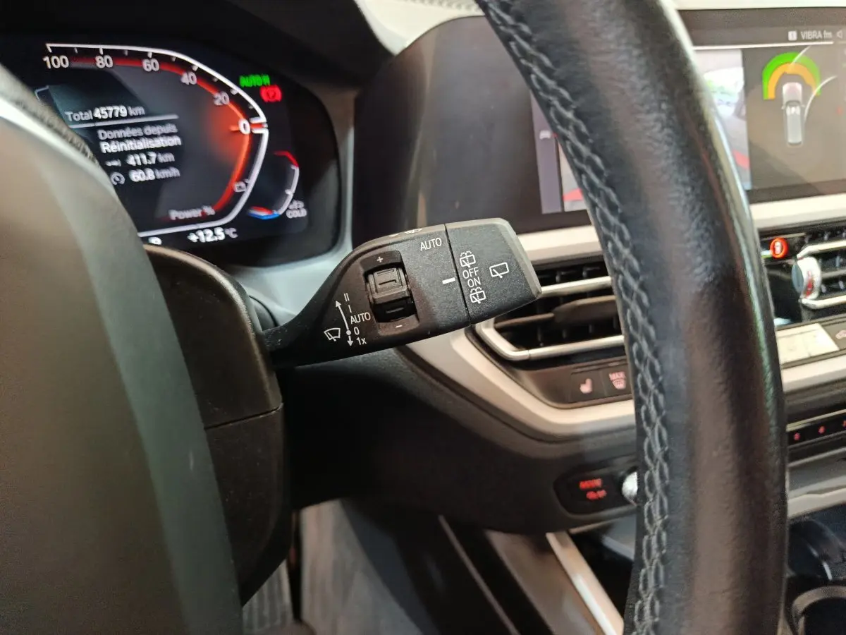 Gros plan sur le commodo de lève-glace à gauche du volant cuir d'une BMW Série 3 gris clair, tableau de bord numérique visible.