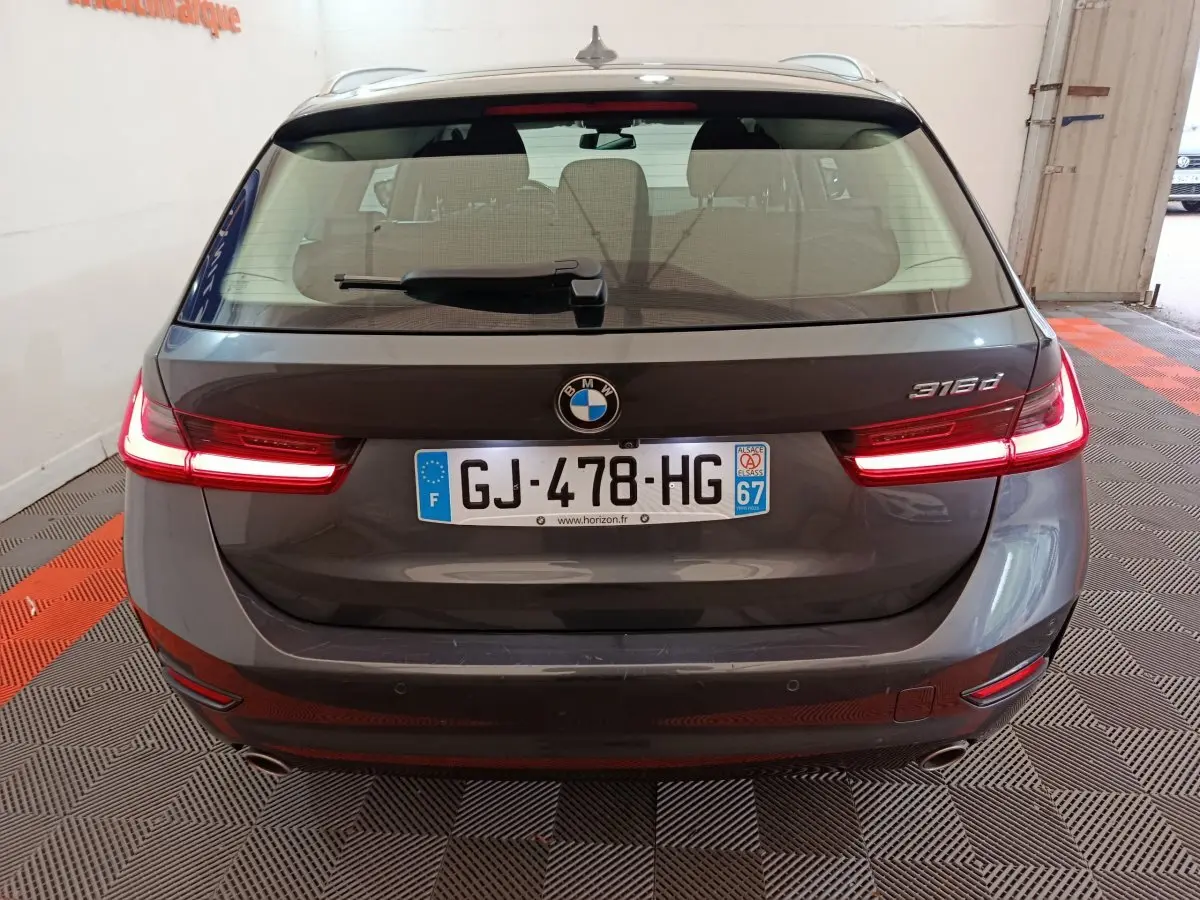 Vue arrière d'une BMW Série 3 316d gris clair avec feux LED allumés et plaque d'immatriculation française visible.