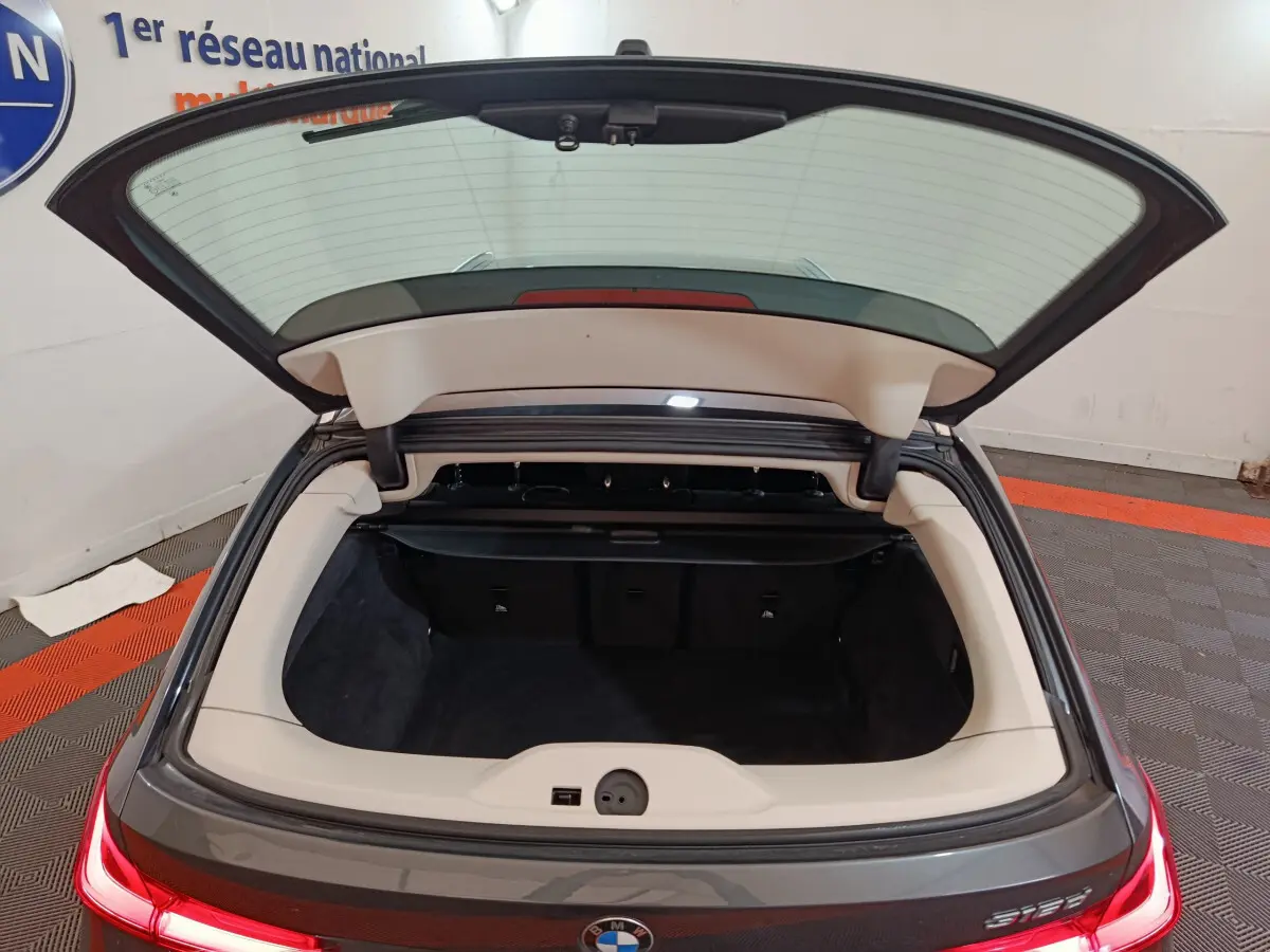 Coffre ouvert vue arrière d'une BMW Série 3 316d gris clair, feux arrière LED allumés dans un garage.