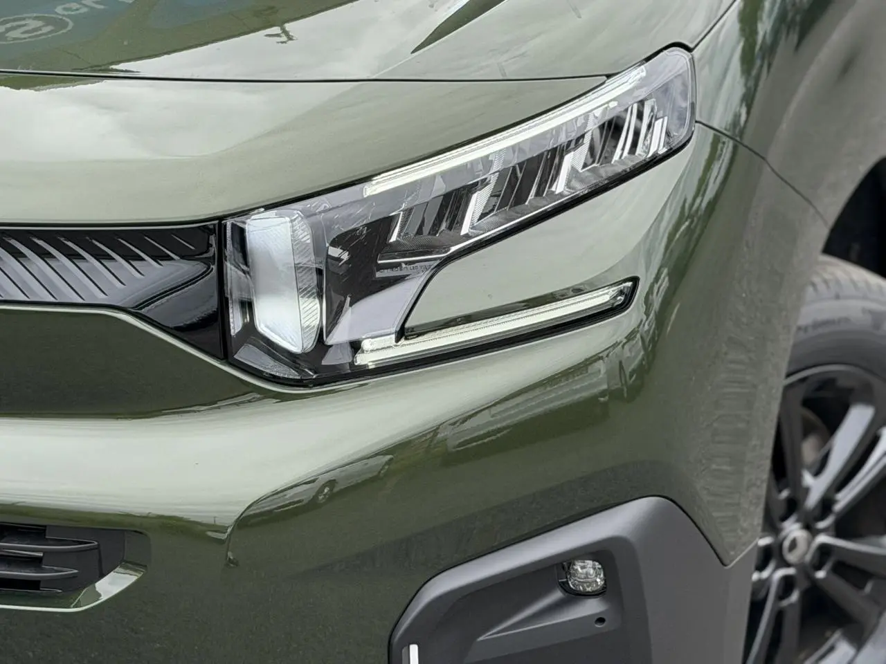 Gros plan sur le phare avant droit du Citroën Berlingo Multispace vert Sirkka avec jante alliage noire visible.