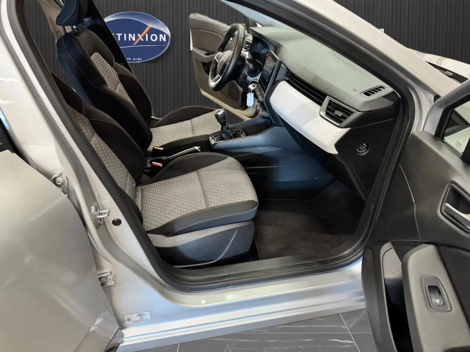 Vue intérieure côté conducteur de la Renault Clio V gris aluminium, montrant les sièges tissu bicolore et le tableau de bord moderne.