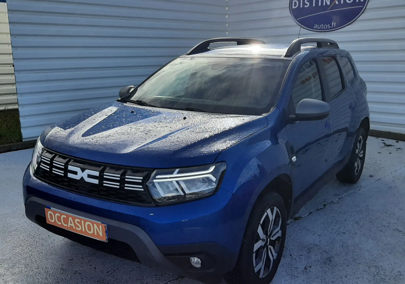 Dacia Duster bleu 2023 en 3/4 avant droit, avec toit ouvrant et jantes bi-ton visibles, stationné devant un mur blanc.