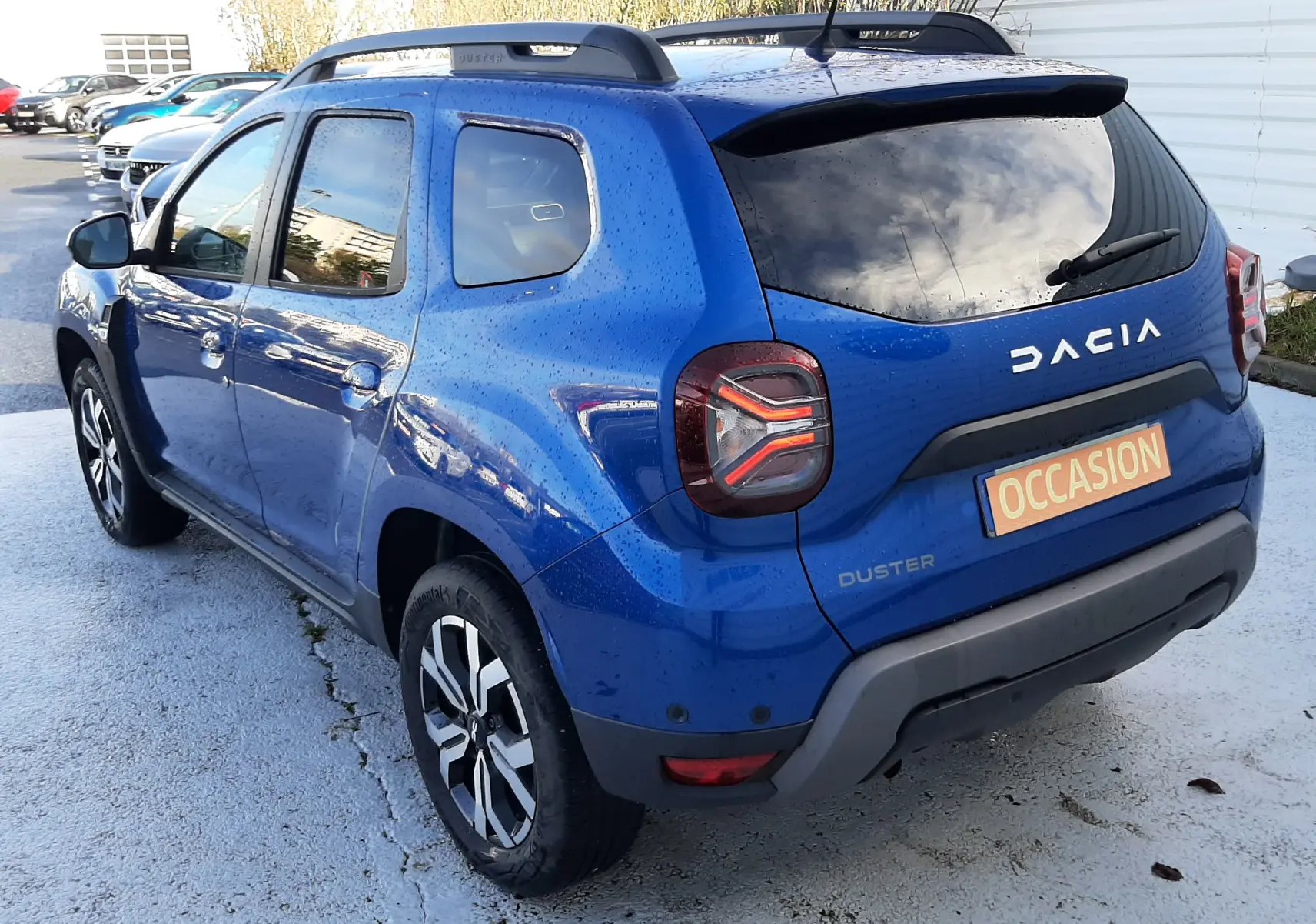 Vue 3/4 arrière droite d'un Dacia Duster bleu 2023 avec toit noir, barres de toit et feux arrière LED distinctifs.