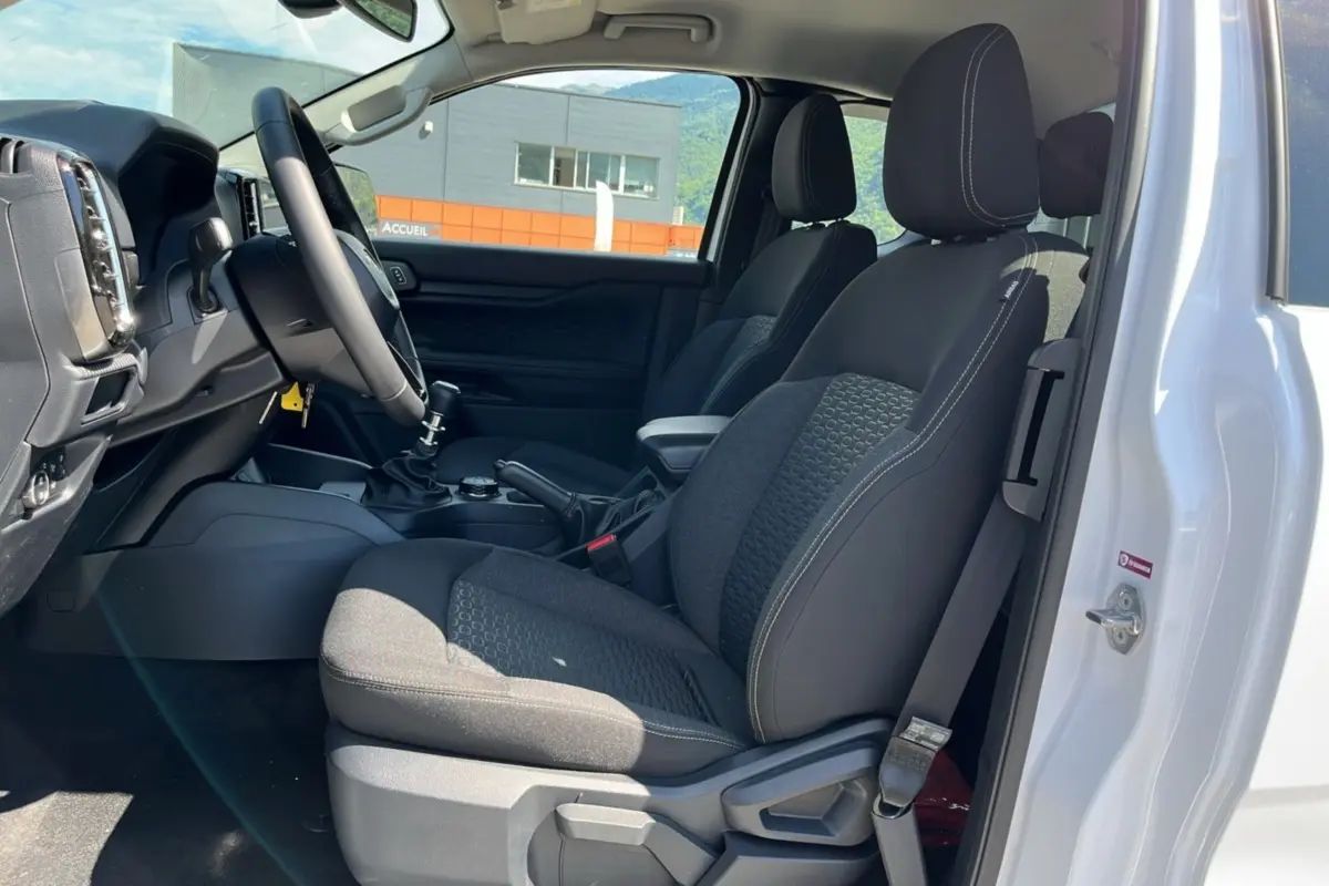 Vue intérieure côté conducteur du Ford Ranger 2025 blanc, montrant sièges tissu noirs et tableau de bord avec volant multifonctions.