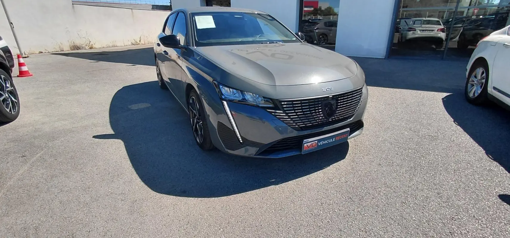 Peugeot 308 gris vue 3/4 avant droit, avec calandre noire distinctive et feux LED verticaux allumés.