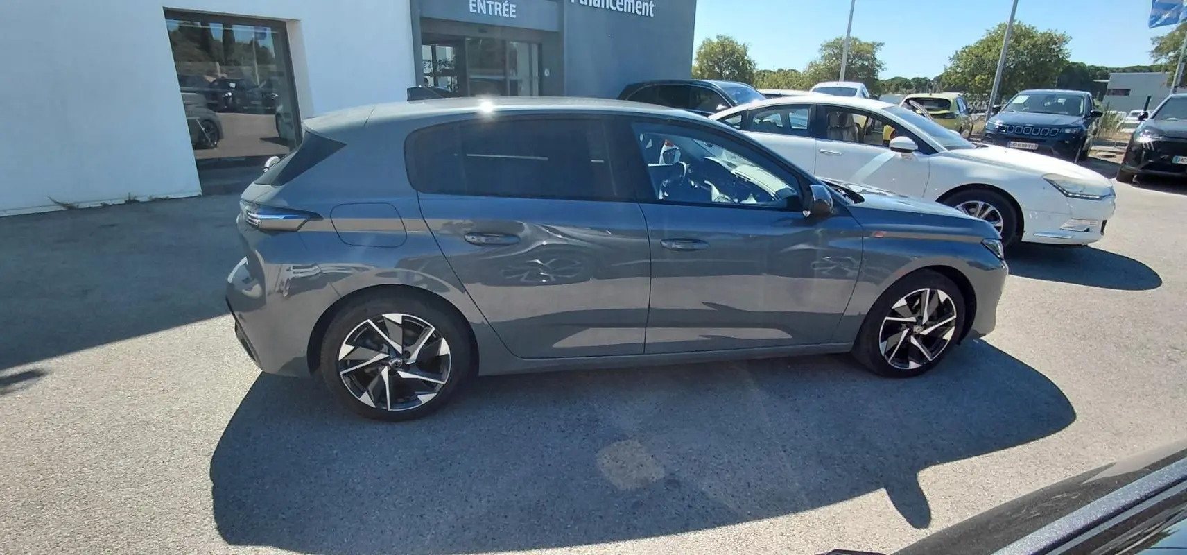 Peugeot 308 gris vue de profil côté gauche, stationnée en extérieur devant un bâtiment blanc.
