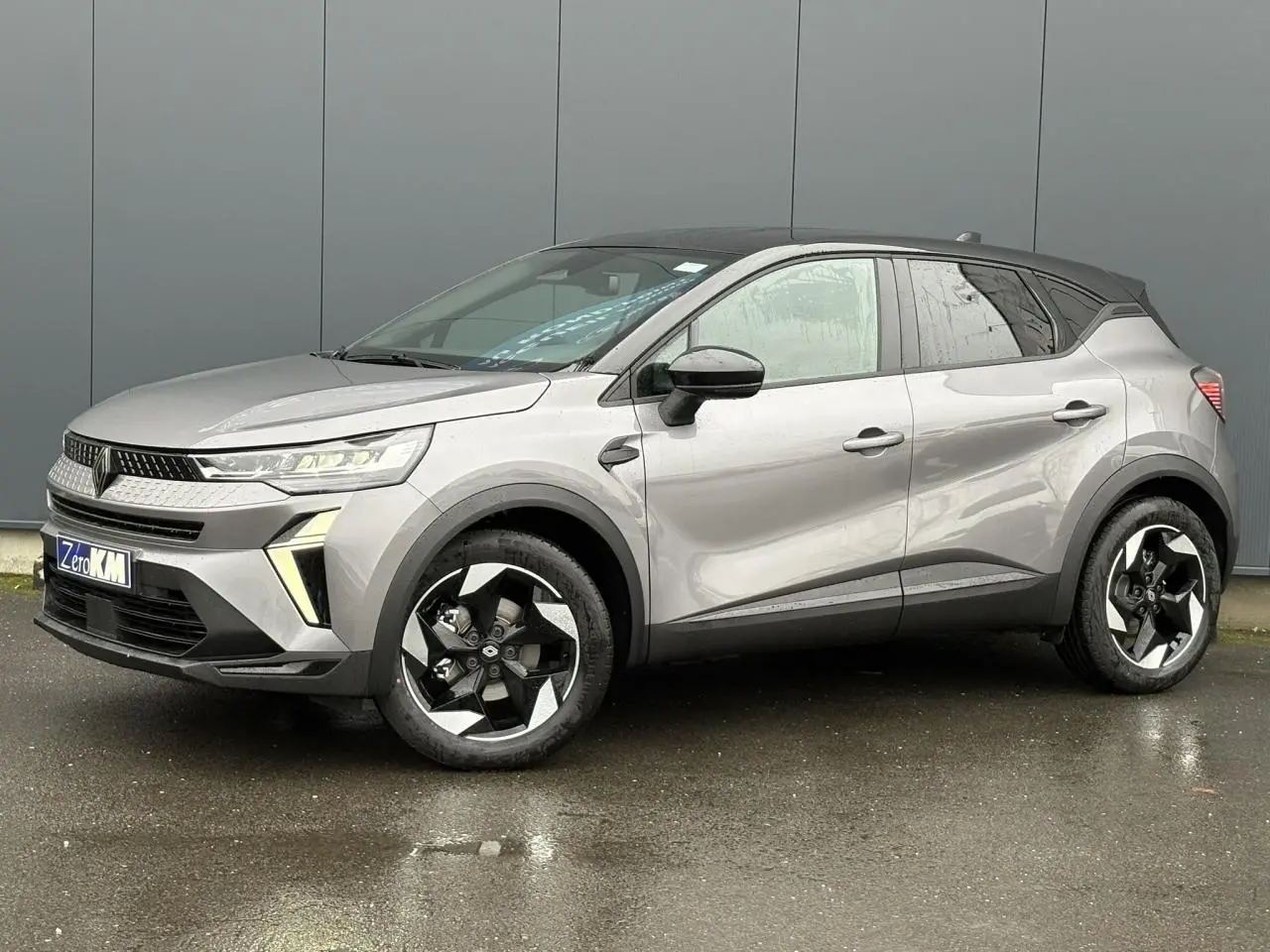 Renault Captur 2025 gris Cassiopée avec toit noir, vue 3/4 avant droit sur sol humide, jantes noires distinctives.