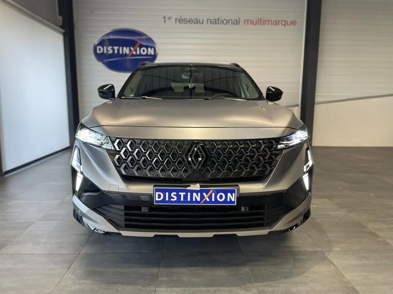 Vue frontale d'un Renault Austral gris satin mat avec calandre noire et feux LED allumés en intérieur showroom.