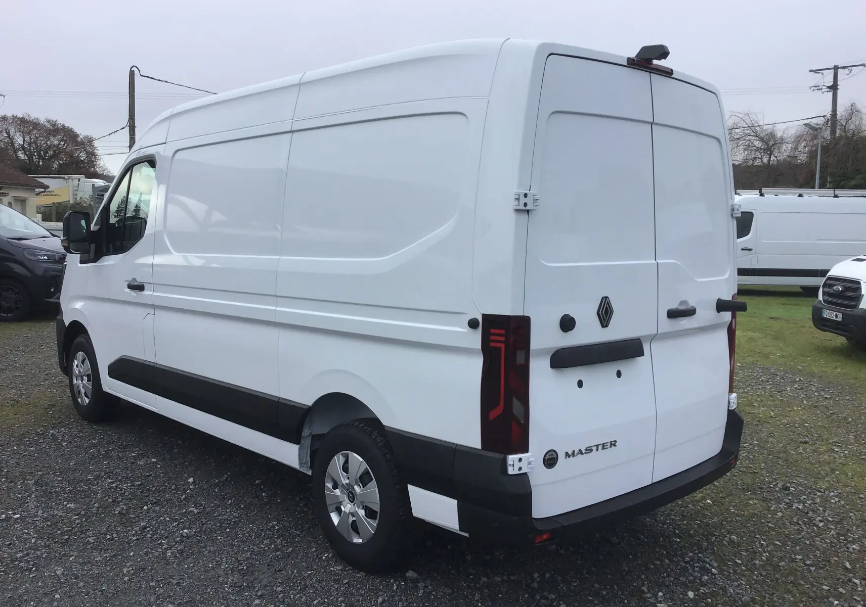 Vue 3/4 arrière droite d’un Renault Master blanc L2H2 avec feux arrière verticaux et porte arrière fermée sur un sol gravillonné.