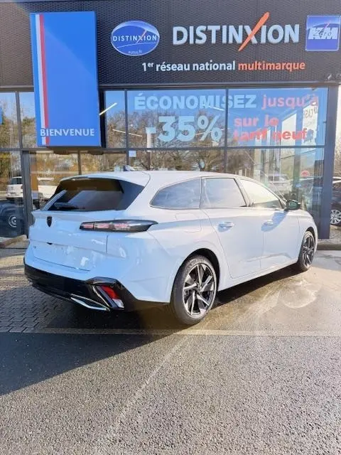 Vue 3/4 arrière droite d'une Peugeot 308 Hybrid 145 e-DCS6 Allure blanche garée devant un concessionnaire multimarque.