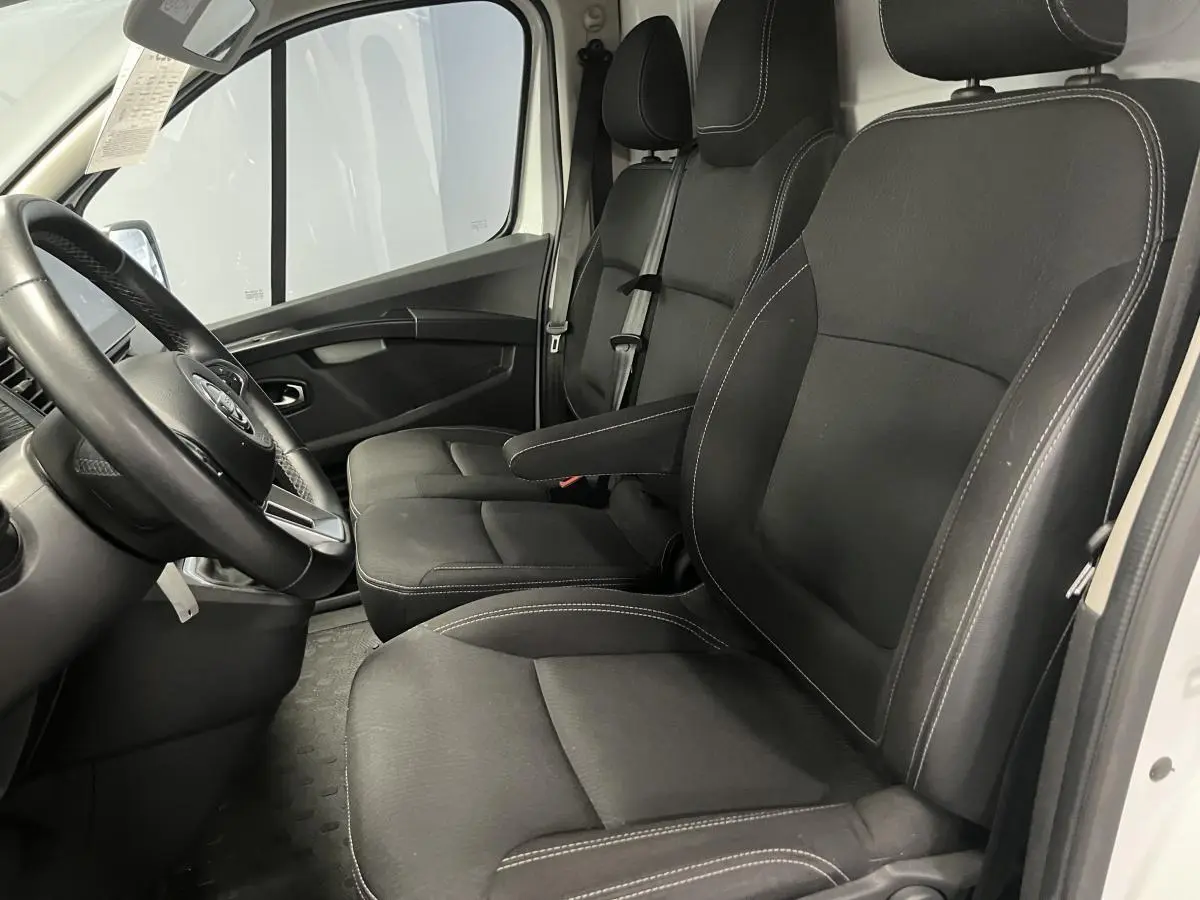 Vue intérieure côté conducteur du Nissan Primastar blanc 2022, sièges tissu gris avec accoudoir et volant simili-cuir.