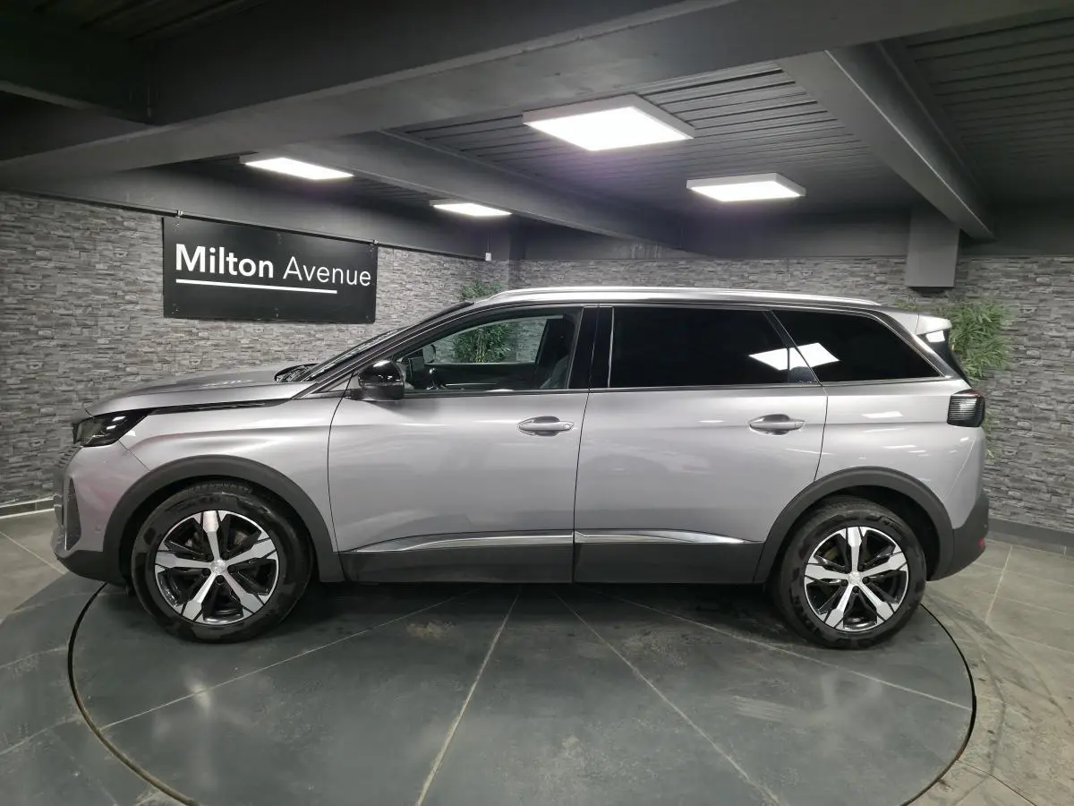 Profil côté gauche du Peugeot 5008 gris métallisé 2022 avec jantes alliage bi-ton et vitres surteintées dans un showroom.