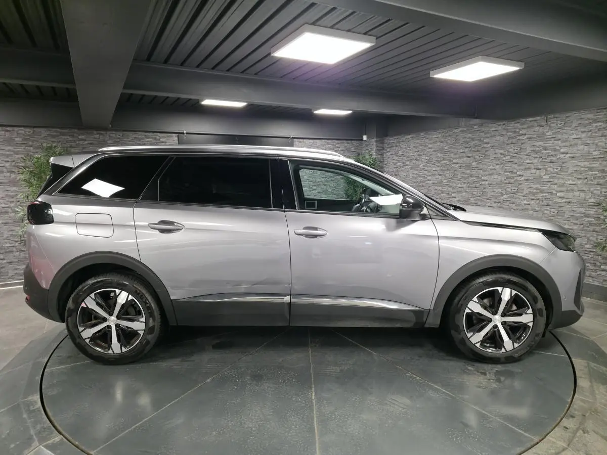 Profil côté gauche d'un Peugeot 5008 gris métallisé avec jantes alliage bi-ton et vitres surteintées en intérieur showroom.